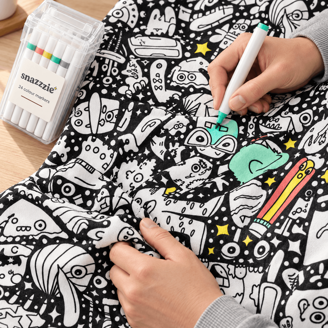 A hand colouring the Doodle Club print beside a case of markers