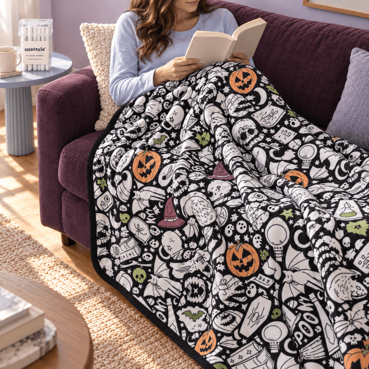 Woman reading on a plum sofa under a partly coloured Spooky Season blanket, with the marker case on a side table